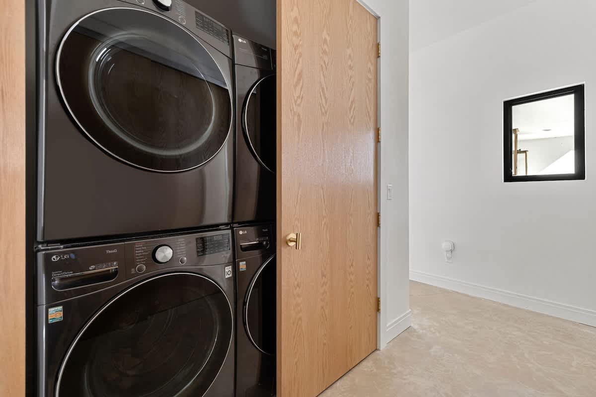 A dedicated laundry area featuring a modern black stacked washer and dryer unit, neatly tucked into an alcove next to a light wooden door, with white walls and light-colored floor tiles.