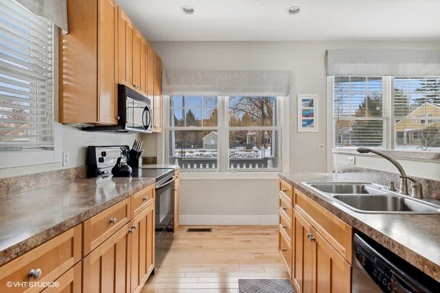Bright kitchen with light wood cabinetry, dark speckled countertops, and stainless steel appliances, featuring a double sink under a large window overlooking a snowy outdoor scene.