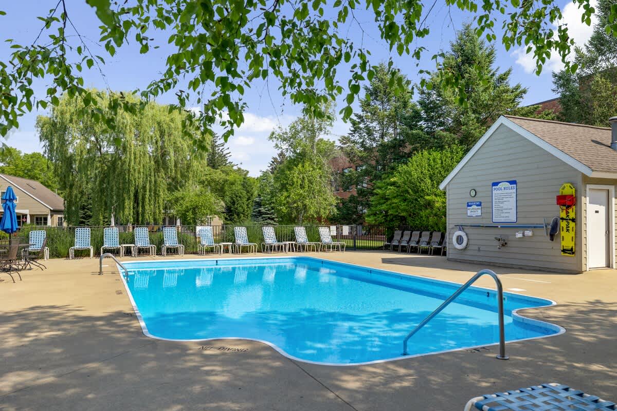 A sparkling blue rectangular swimming pool is surrounded by a concrete deck with numerous lounge chairs and umbrellas. A small pool house with "POOL RULES" signage stands to the right, all set amidst green trees.