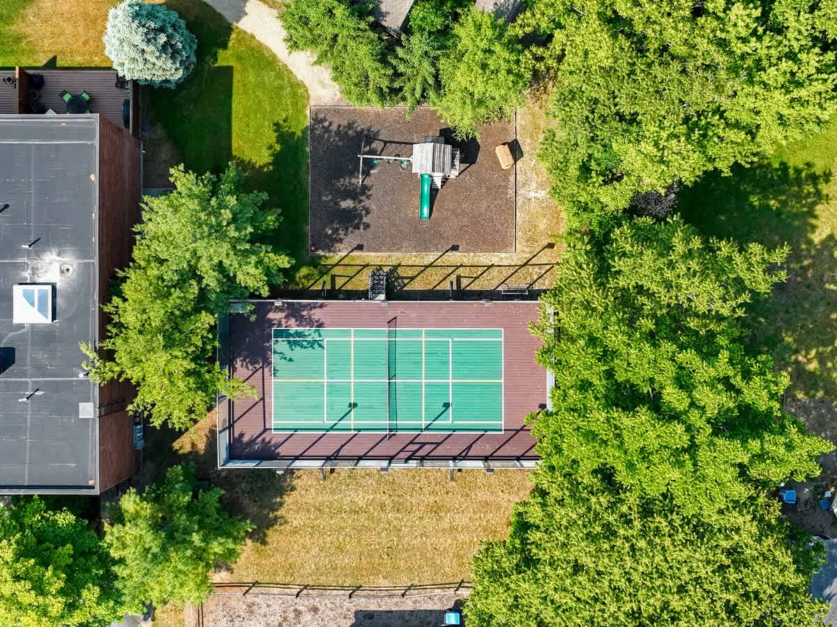 An aerial perspective reveals a green tennis court enclosed by a fence, adjacent to a children's playground with swings and a slide. Both are surrounded by dense green trees.