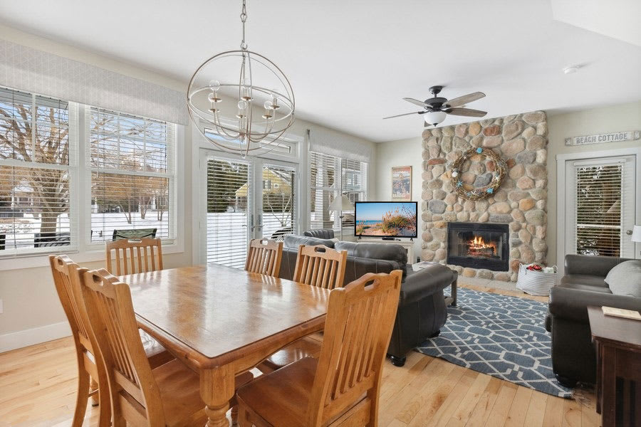 Open-concept living and dining area with a wooden dining table and chairs under a modern spherical chandelier, adjacent to a living space with a stone fireplace and dark leather sofas, all with views of a snowy landscape through large windows.