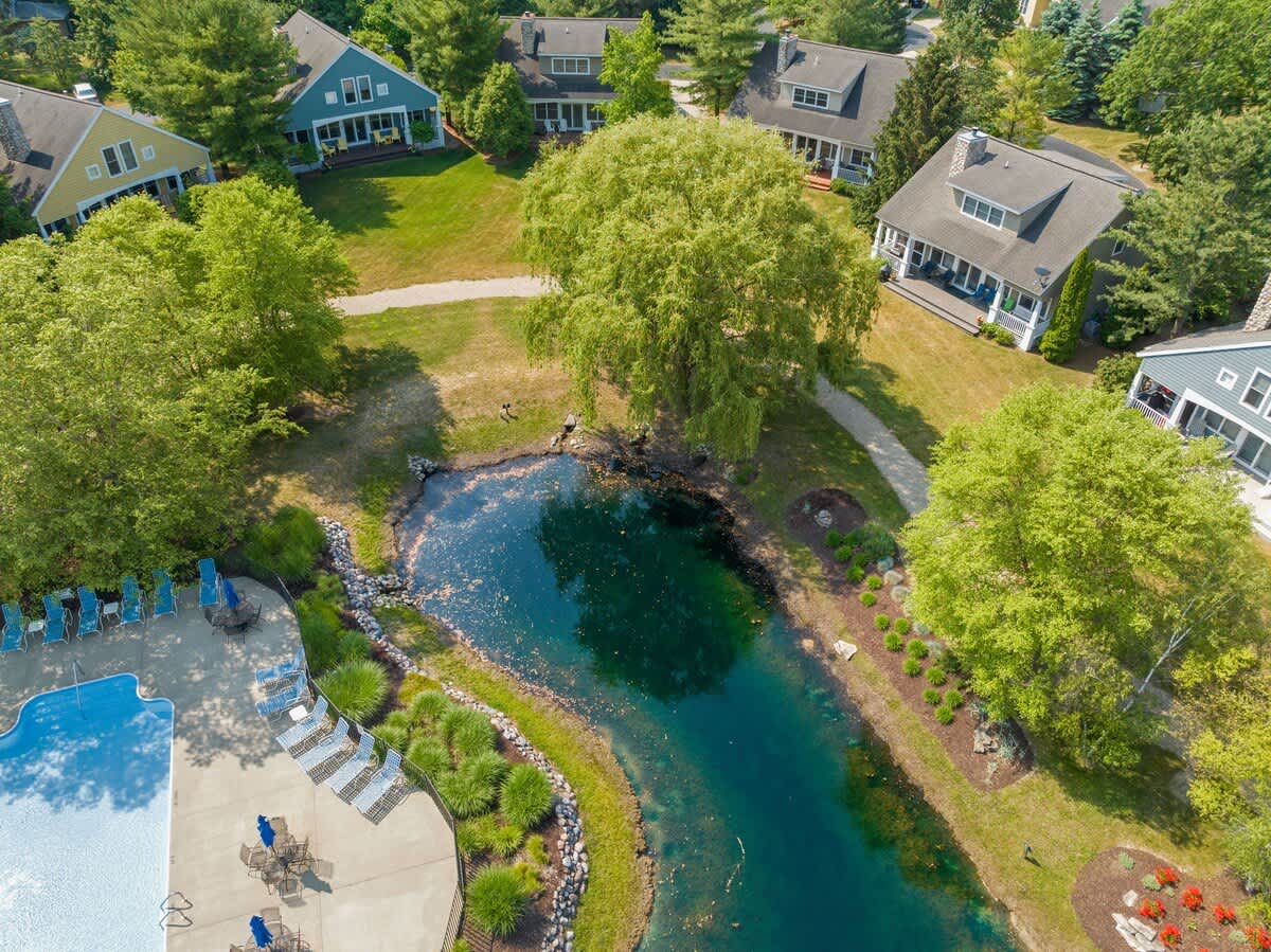 A high-angle aerial shot captures a scenic community with a dark blue pond, a bright swimming pool, and several residential homes nestled among abundant green trees and landscaped grounds.