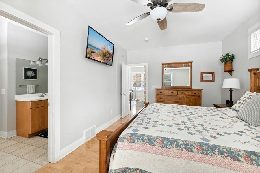 Master bedroom with light wood flooring and light gray walls, featuring a wooden bed frame with a colorful quilt, a wooden dresser with a mirror, and a wall-mounted TV, with an en-suite bathroom visible through an open doorway.