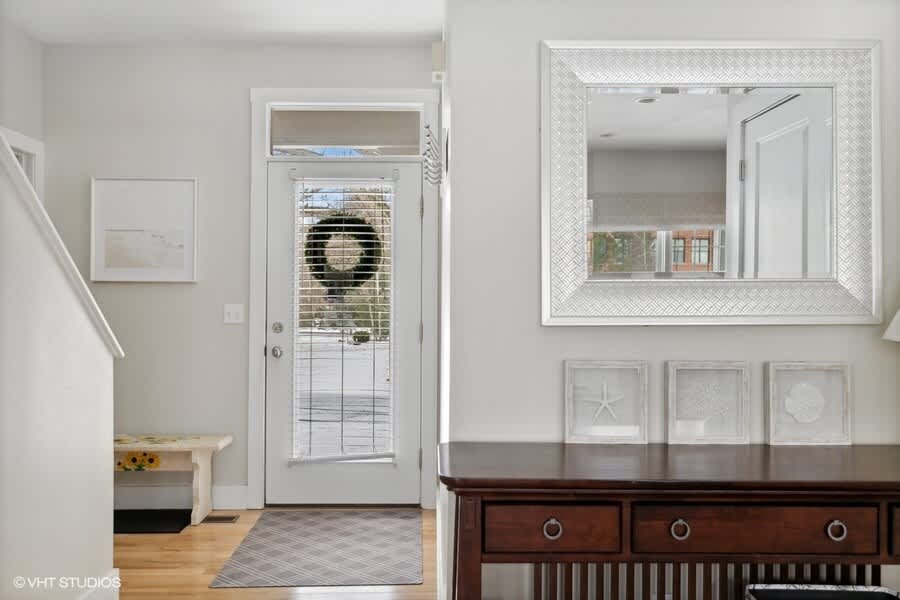 Inviting entryway with light hardwood floors, a white front door adorned with a wreath, a large decorative mirror above a dark wooden console table, and a small white bench, creating a welcoming first impression.