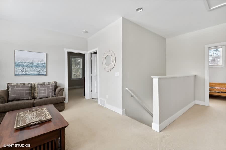 Upper-level landing area with light gray walls and beige carpet, featuring a small sofa and coffee table, leading to a hallway and stairs, with a round decorative mirror on the wall.