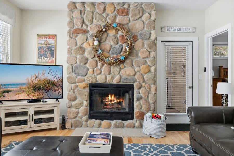 Cozy living room featuring a large stone fireplace with a decorative wreath, a flat-screen TV on a white media console, and a dark leather sofa, all set on light hardwood floors with a blue and white patterned rug.