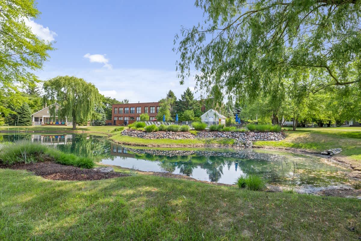 A serene pond reflects the blue sky and surrounding greenery, with a large brick building and a community pool area visible in the distance. Lush trees, including a weeping willow, frame the tranquil landscape.
