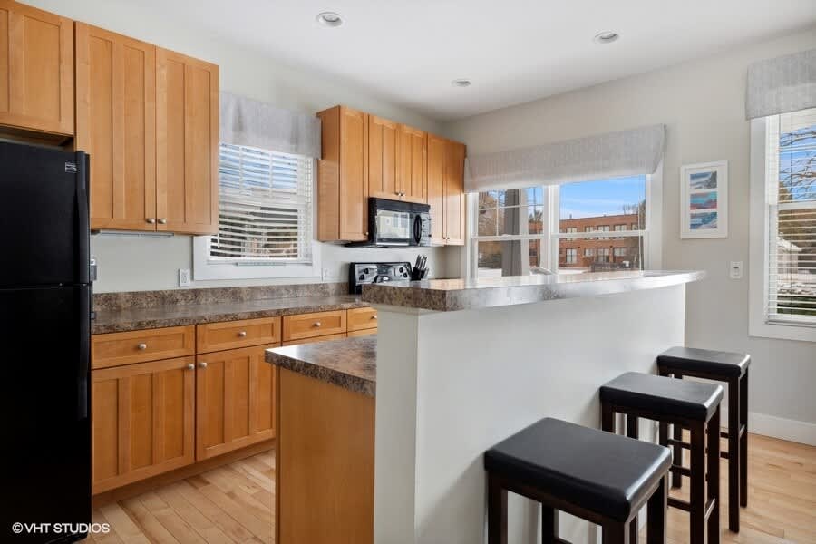 Functional kitchen featuring light wood cabinets, dark speckled countertops, a black refrigerator, and a breakfast bar with three dark stools, illuminated by natural light from multiple windows.