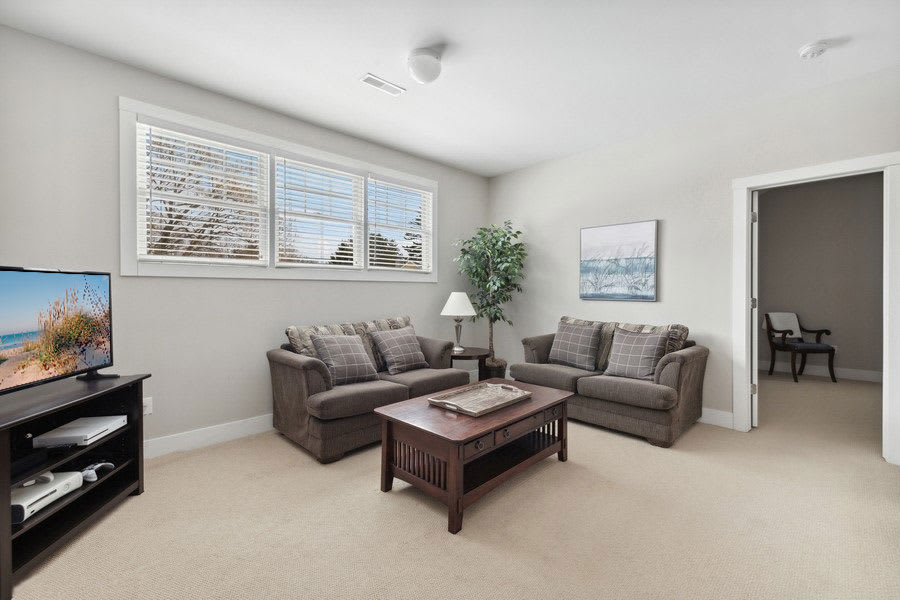 Secondary living space or loft area with light gray walls and beige carpet, furnished with two brown sofas, a wooden coffee table, and a flat-screen TV on a dark media console, illuminated by natural light from multiple windows.