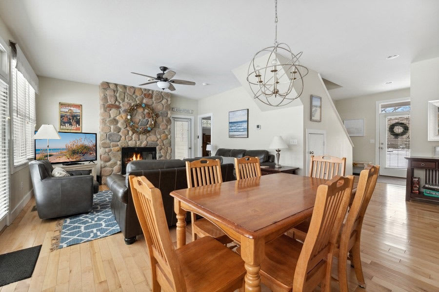 Spacious open-plan living and dining area with light hardwood floors, a wooden dining table, and a comfortable living space featuring dark leather sofas and a prominent stone fireplace, enhanced by a modern chandelier and ceiling fan.