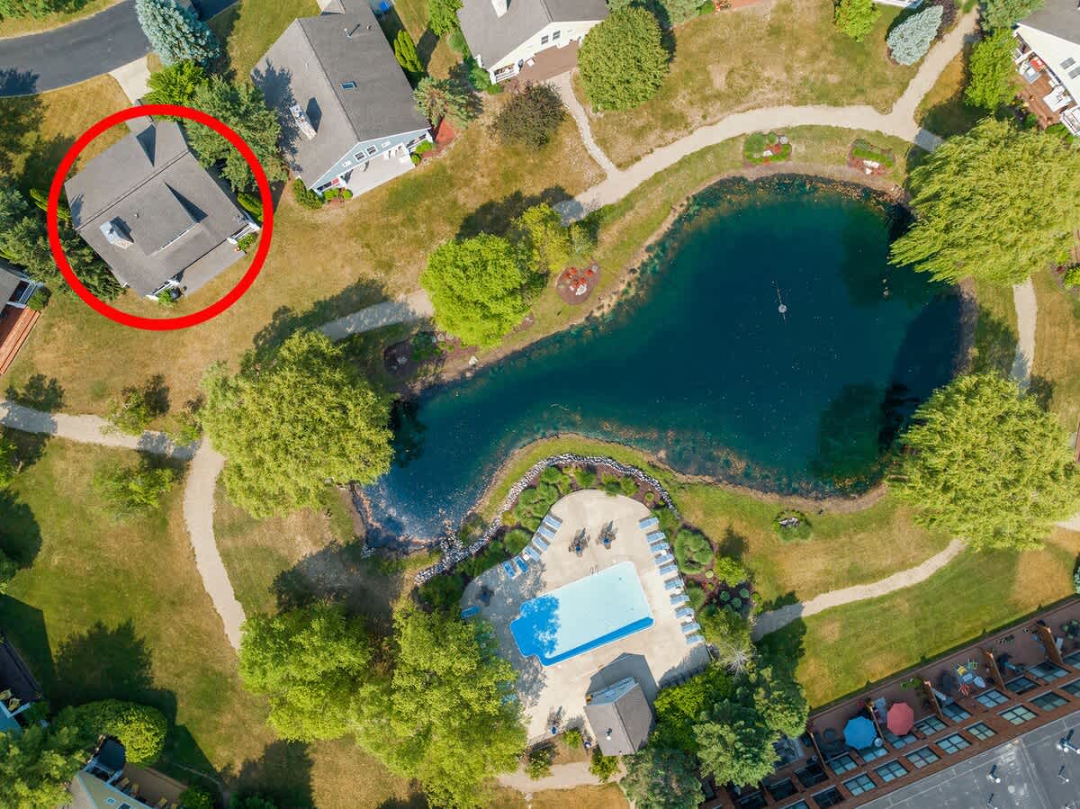An aerial view highlights the property, circled in red, situated near a large, irregularly shaped pond and a community swimming pool. Winding paths connect various houses and green spaces under a clear sky.