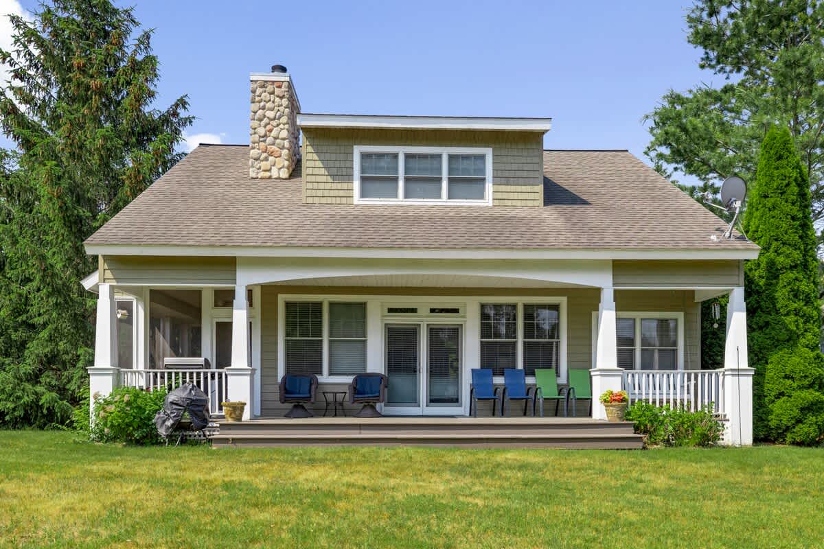 The back of a light green house features a spacious wooden deck with outdoor seating and a grill, leading to a screened porch. A stone chimney rises on the left, and the house is surrounded by a lush green lawn and tall trees.