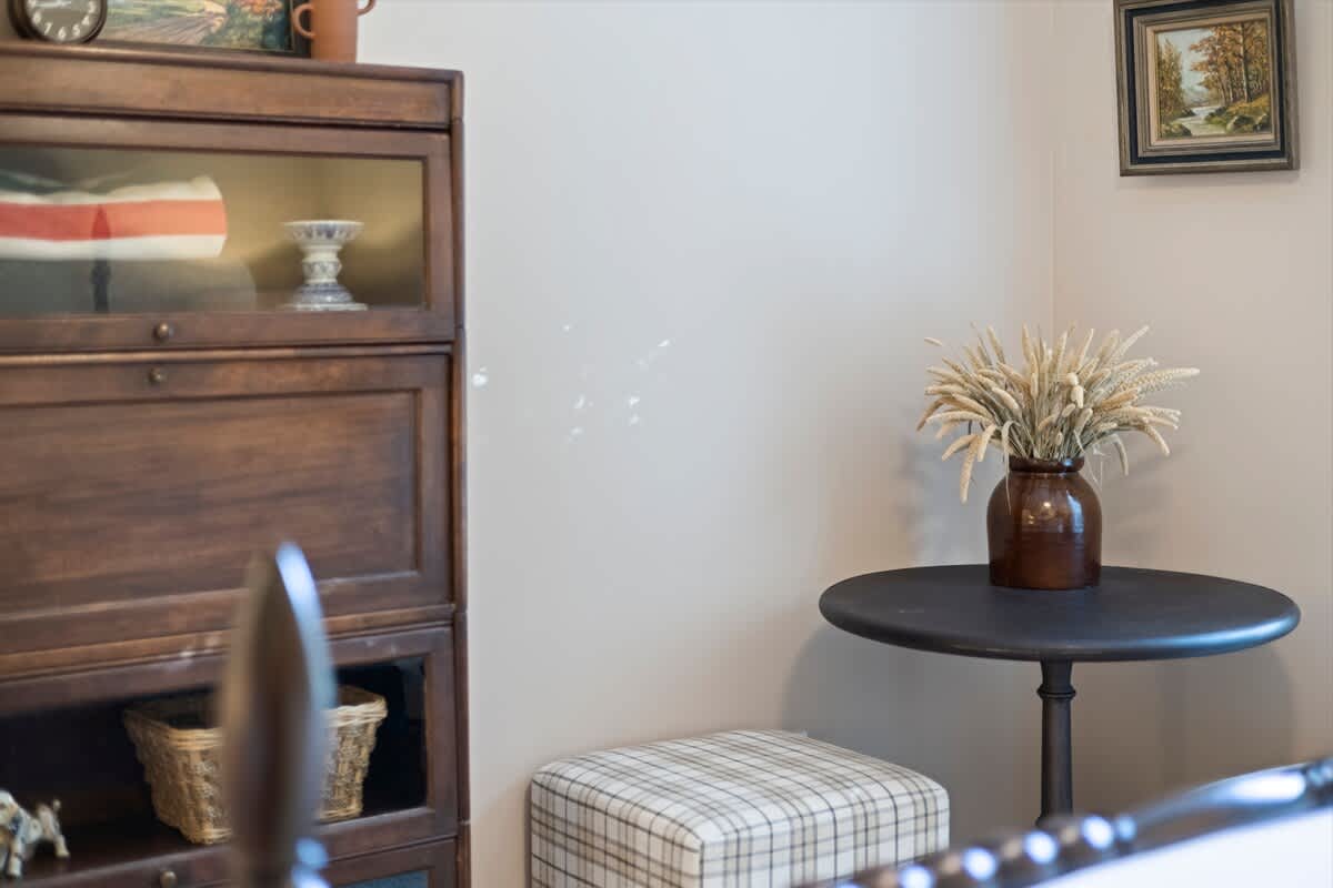 Corner of a bedroom featuring a dark wood cabinet, a small round table with a vase of dried grass, and a plaid ottoman. The wall is light beige, and a framed picture hangs above the table.