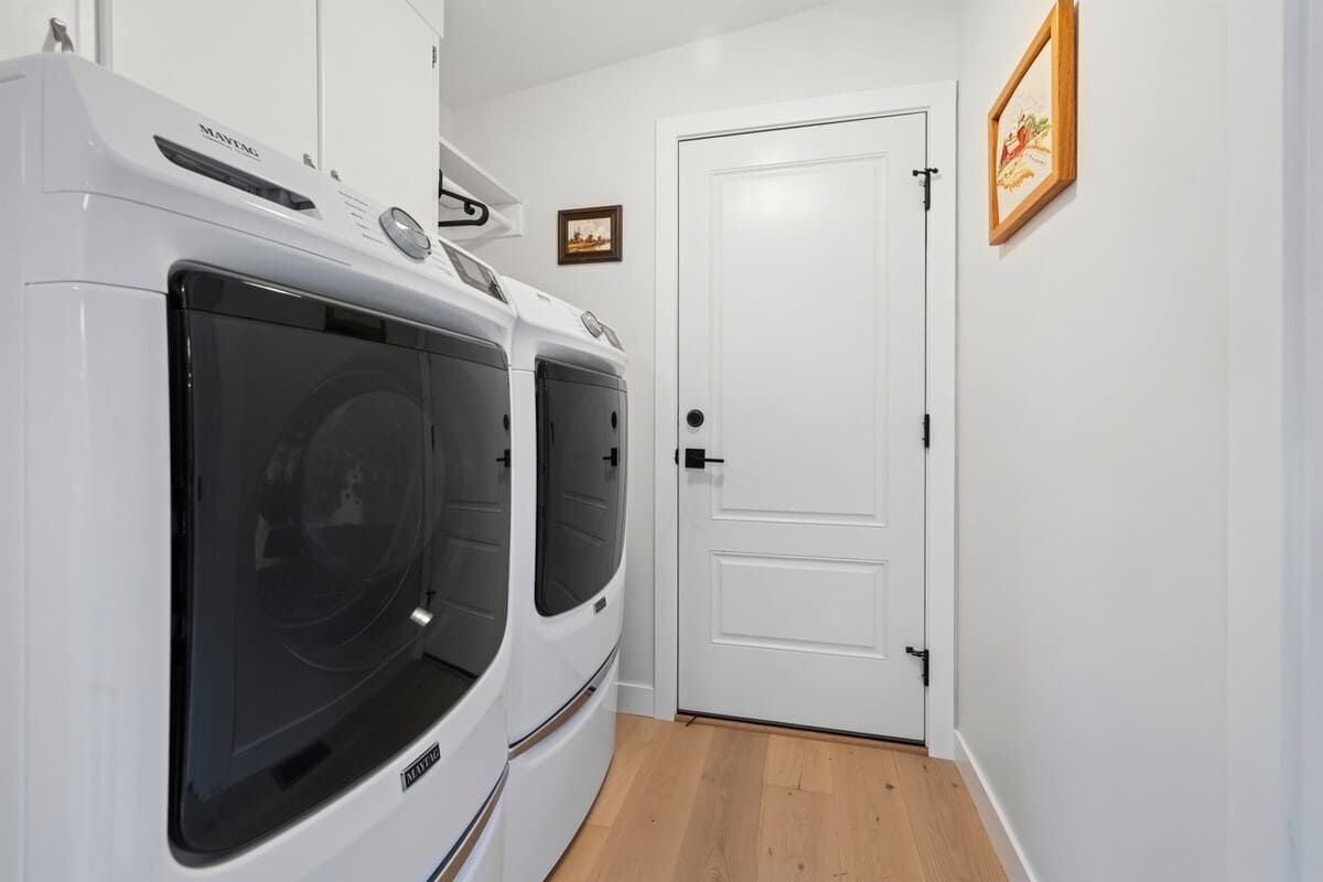 A bright laundry room equipped with a white Maytag washer and dryer, a white door with black hardware, and light hardwood flooring, featuring white walls and a small framed picture.