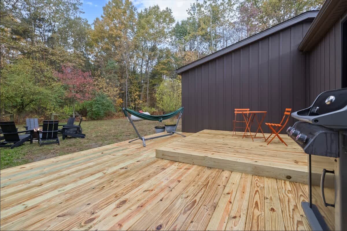 A spacious wooden deck extends from the dark brown house, featuring a BBQ grill, a green hammock, and leading to a grassy yard with a fire pit area and mature trees.