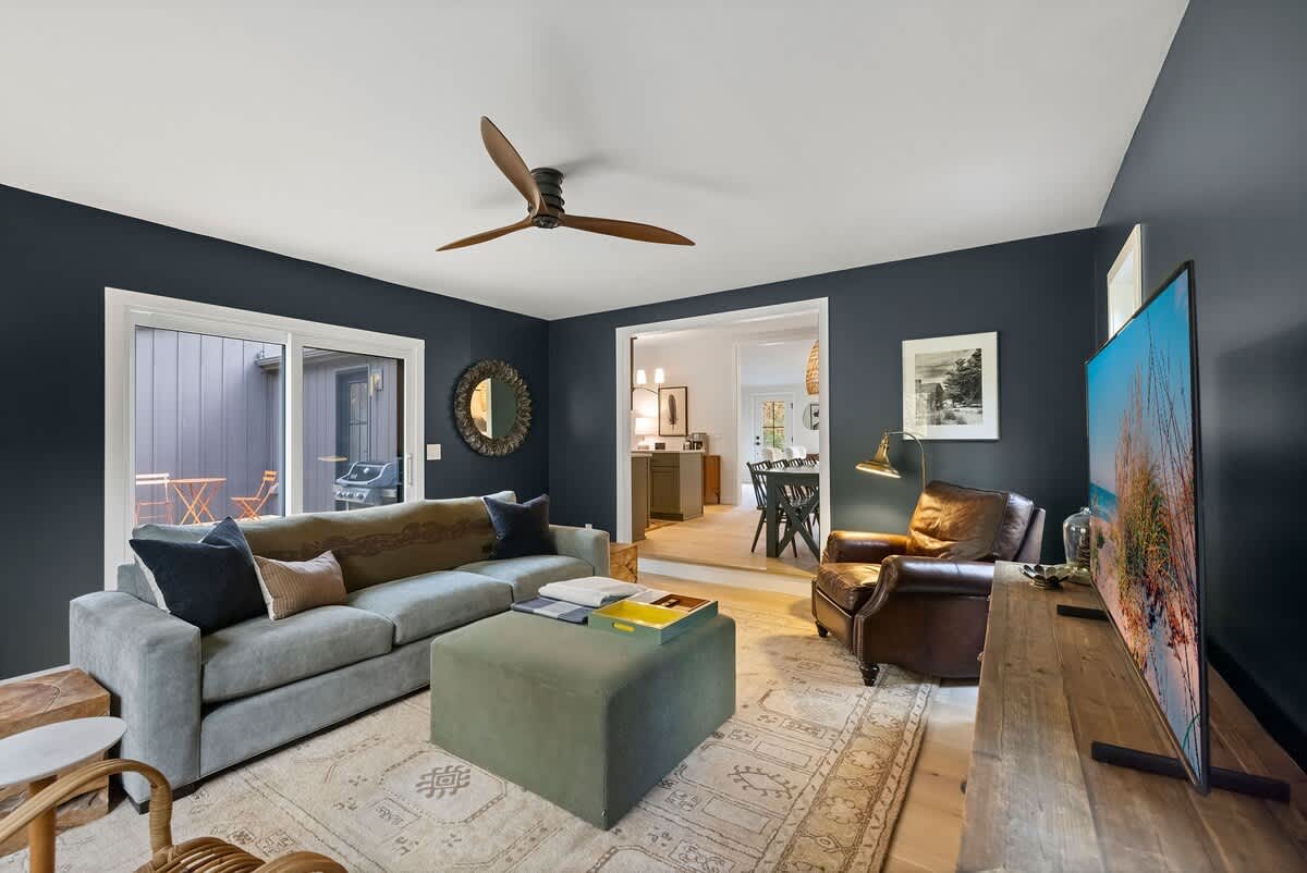 Cozy living room with dark blue walls, a light gray sectional sofa, a leather armchair, and a large flat-screen TV on a rustic wooden console. A ceiling fan with wooden blades is visible overhead.