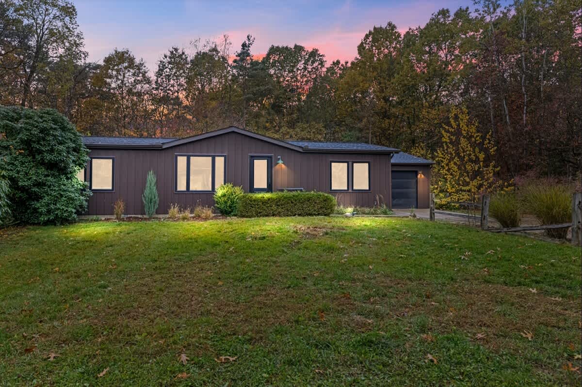 The front exterior of a dark brown single-story house with multiple windows and a front door, surrounded by a green lawn and mature trees under a colorful dusk sky.