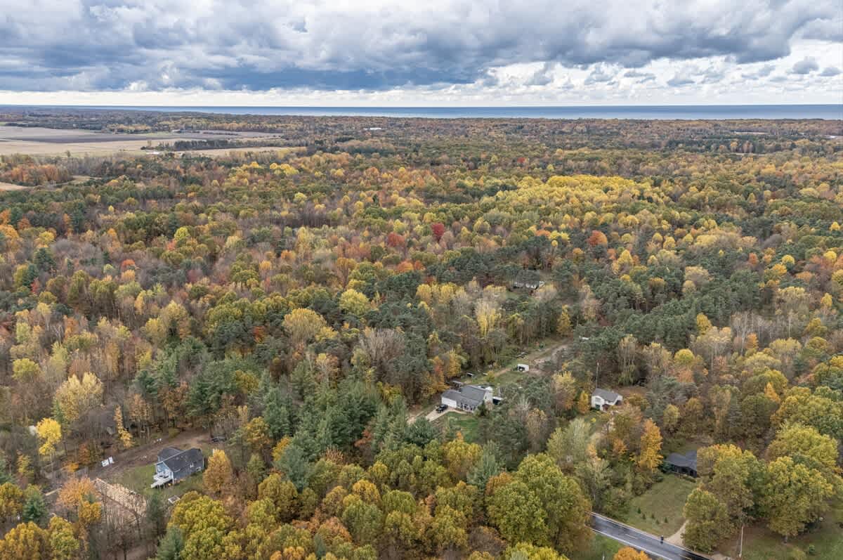 A vast aerial landscape view of the property and surrounding area, dominated by a dense forest of colorful autumn trees, with a large body of water visible on the horizon under a cloudy sky.