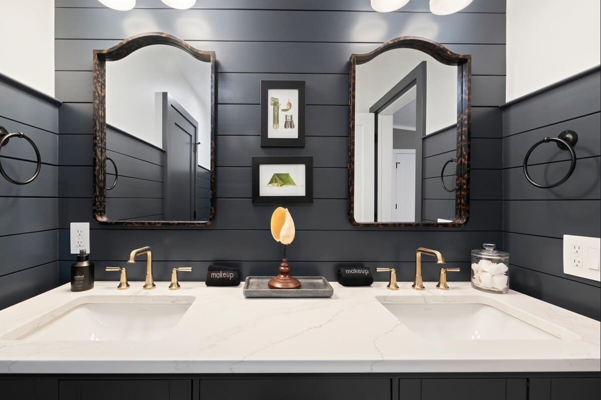 Double vanity bathroom featuring dark shiplap walls, two ornate framed mirrors, brass fixtures, and a white quartz countertop with two undermount sinks. Decorative items include framed art and a large shell.