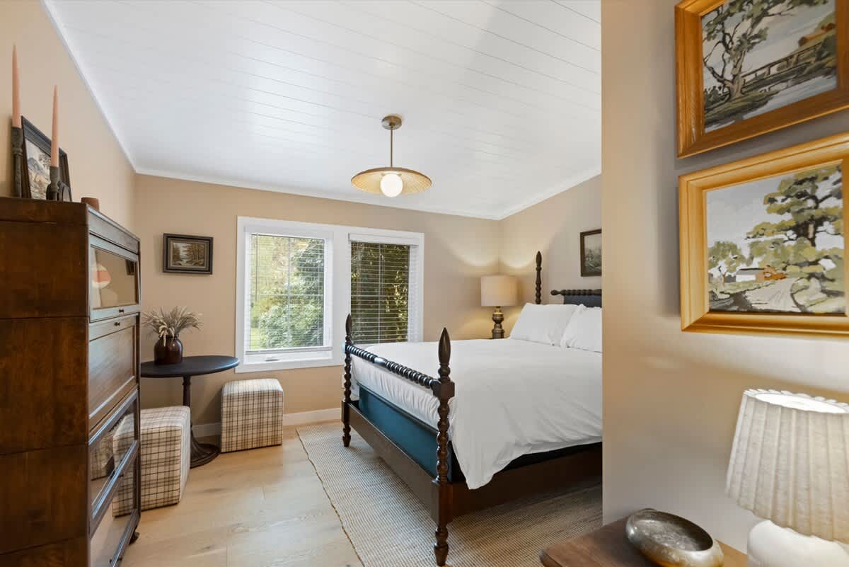 Wider view of a warm bedroom with a dark wood four-poster bed, light wood floors, and light beige walls. The room includes a dark wood dresser, a small round table, and framed artwork.