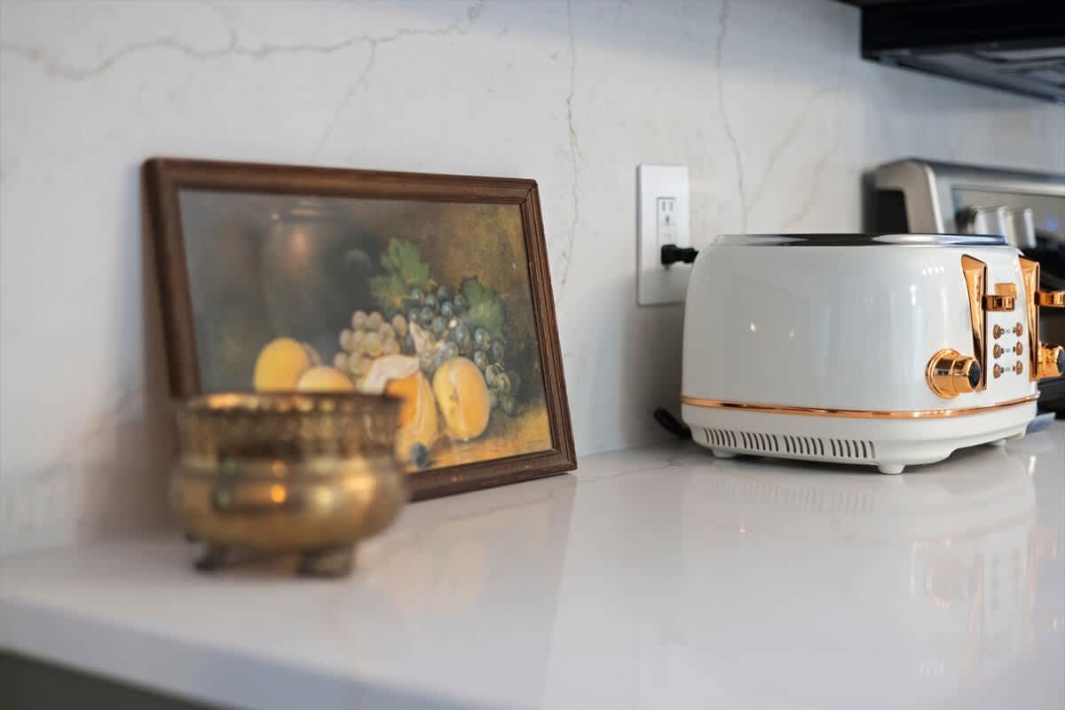 Close-up of a white quartz countertop with a vintage-style white and rose gold toaster. A small framed still-life painting of fruit and a brass bowl add decorative accents to the kitchen space.