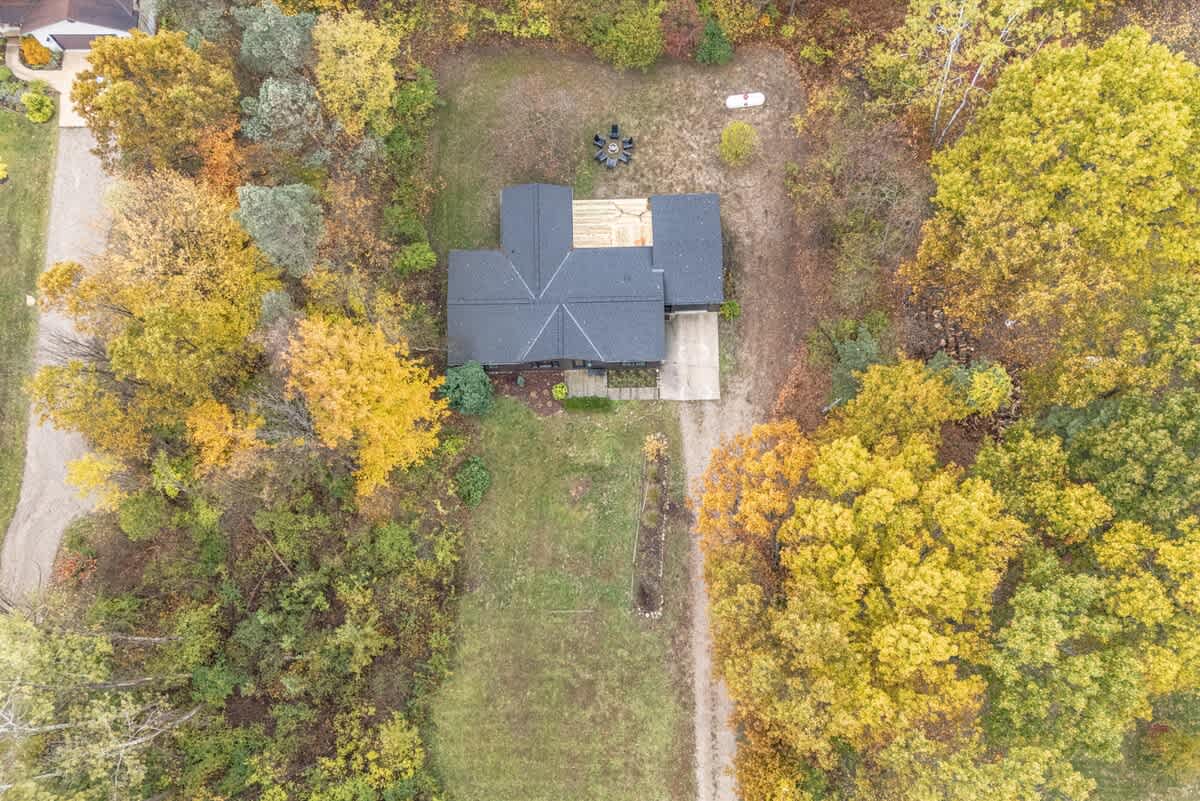 A top-down aerial view of the house with its dark roof, showing the wooden deck and the fire pit area in the backyard, surrounded by a mix of green and autumn-colored trees.