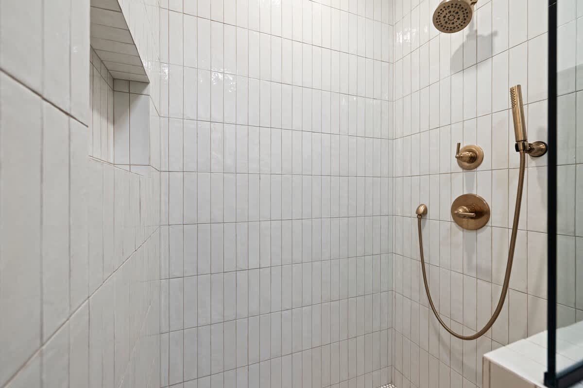 Close-up view inside a walk-in shower featuring white vertical subway tiles, a tiled niche, and modern brass shower fixtures including a rain shower head and handheld sprayer.