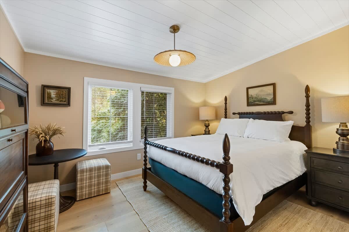 Cozy bedroom with a dark wood four-poster bed, light wood floors, and light beige walls. Two windows with blinds overlook greenery, and a round table with a plaid ottoman sits beside the bed.