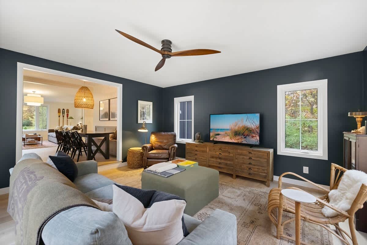 Inviting living room with dark blue walls, a comfortable gray sofa, and a leather armchair, centered around a large area rug. An open doorway leads to a bright dining space.
