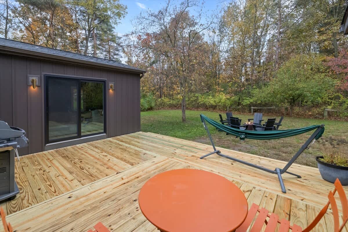 An expansive wooden deck with an orange patio table and chairs, a BBQ grill, and a green hammock, overlooking a grassy backyard with a fire pit area and surrounding autumn trees.