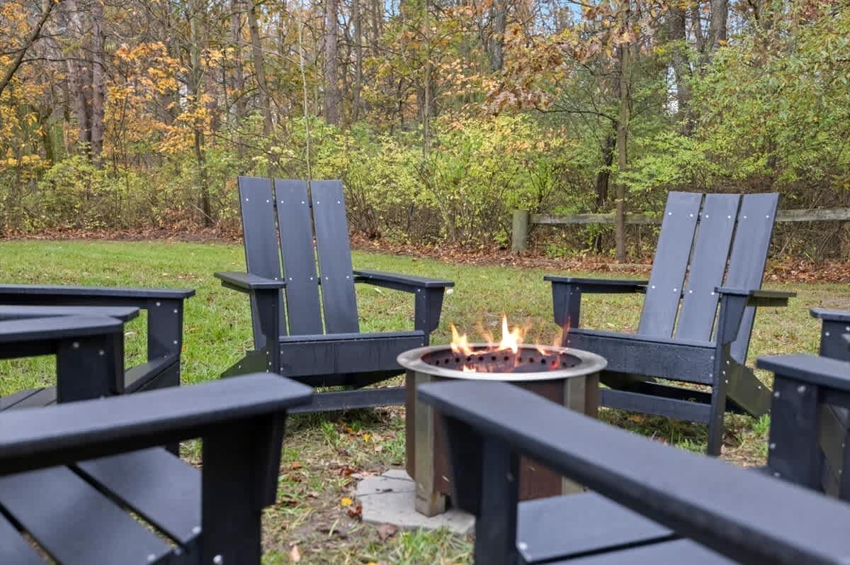 A cozy outdoor fire pit with flames burning, surrounded by several dark Adirondack-style chairs on a grassy patch, with lush autumn trees in the background.