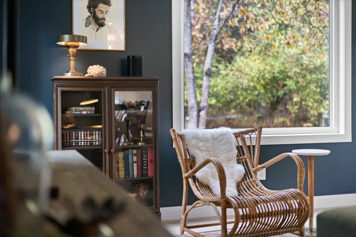 Close-up of a cozy reading nook in the living room, featuring a rattan armchair with a white sheepskin throw, a glass-front display cabinet filled with books, and a window overlooking trees.