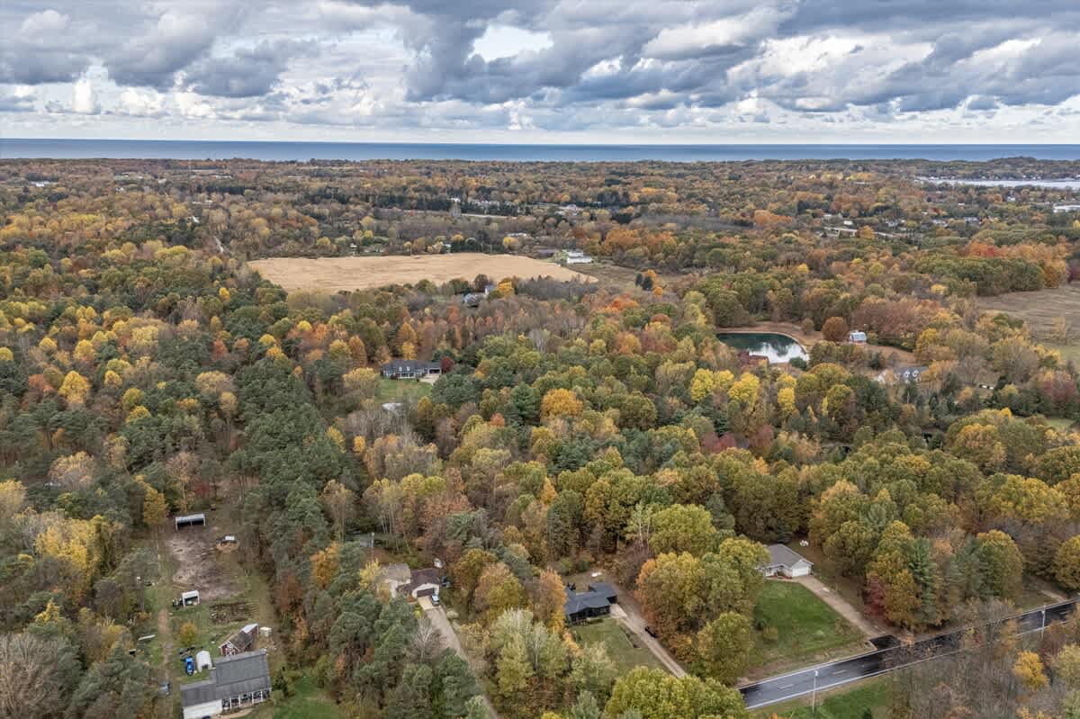 Aerial view of a vast, colorful autumn landscape featuring dense forests, scattered homes, a small pond, and a distant large body of water under a cloudy sky. The scene highlights the property's secluded, natural setting.