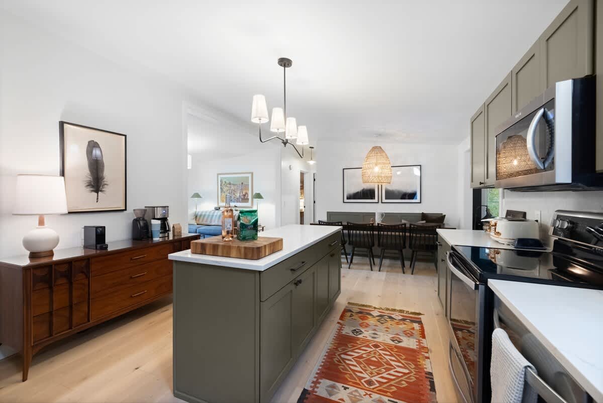 View of the kitchen island with olive green cabinetry and white countertops, extending towards a dining area with a dark table and chairs. A patterned rug adds warmth to the light hardwood floors.