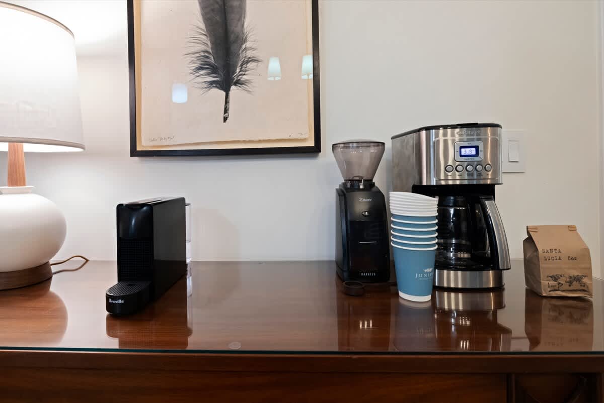 A dedicated coffee station on a polished wooden console table, featuring a Nespresso machine, a coffee grinder, and a drip coffee maker. A framed feather artwork hangs above, with disposable cups and a bag of coffee beans nearby.