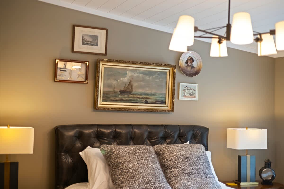 Serene bedroom with light taupe walls and a dark tufted leather headboard. A gallery wall above the bed features various framed artworks, including a prominent seascape painting.