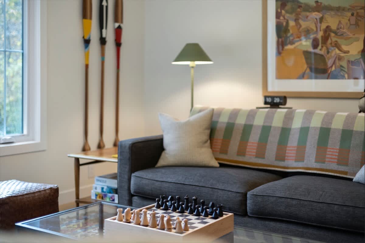 Close-up of a dark gray sofa adorned with a plaid blanket and throw pillows, positioned next to a window with colorful decorative oars. A glass coffee table holds a wooden chess set, with a green-shaded floor lamp providing ambient light.