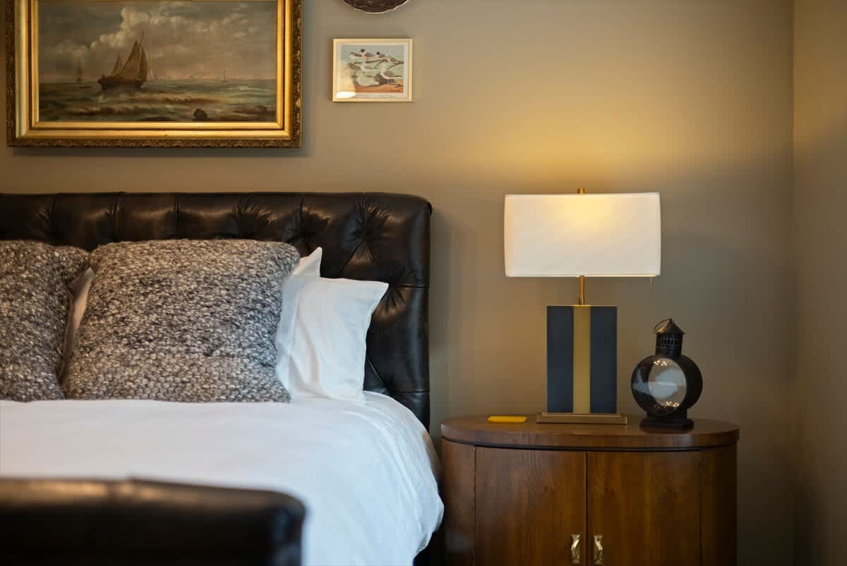 Detailed view of the bedroom's bedside area, showcasing a dark tufted leather headboard, a round wooden nightstand, and a modern table lamp with a rectangular base.
