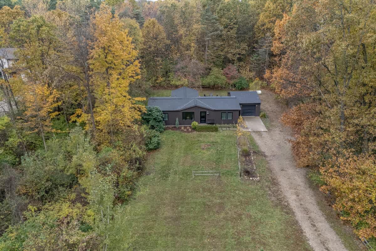 An aerial view showcasing the dark-roofed house nestled among vibrant autumn trees, with a long gravel driveway leading up to it and a large grassy yard.
