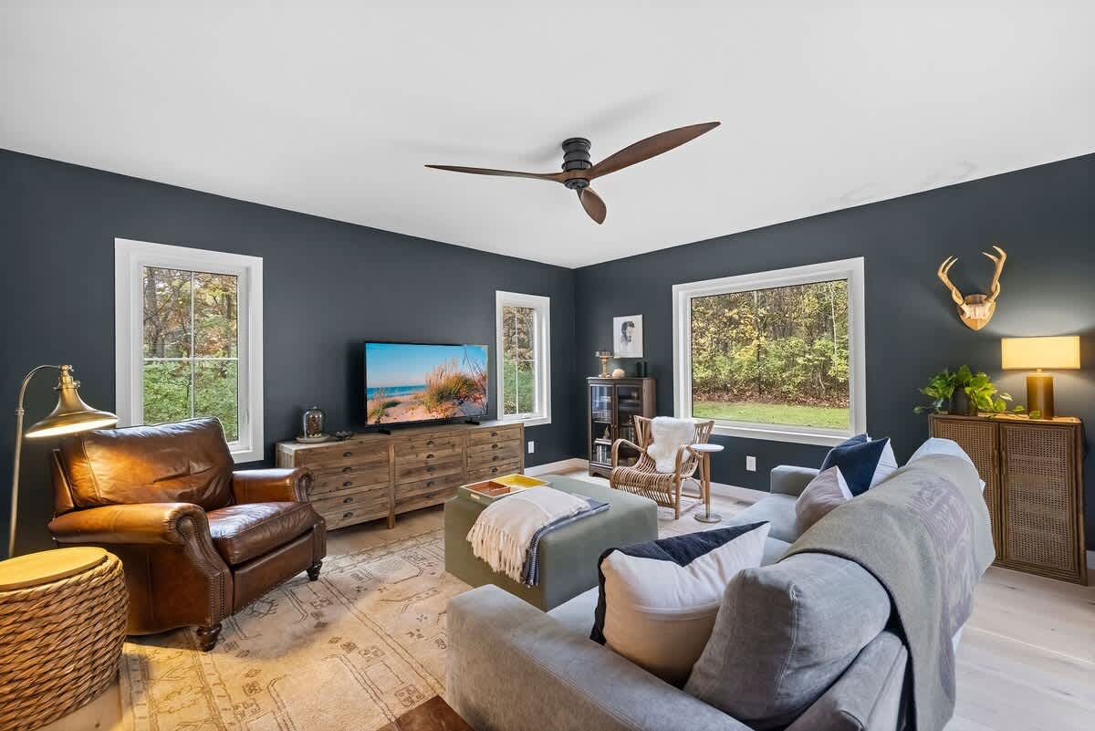 Spacious living room with dark blue walls and light hardwood floors, featuring a gray sofa, a leather armchair, and a large TV. Multiple windows offer views of the surrounding greenery.