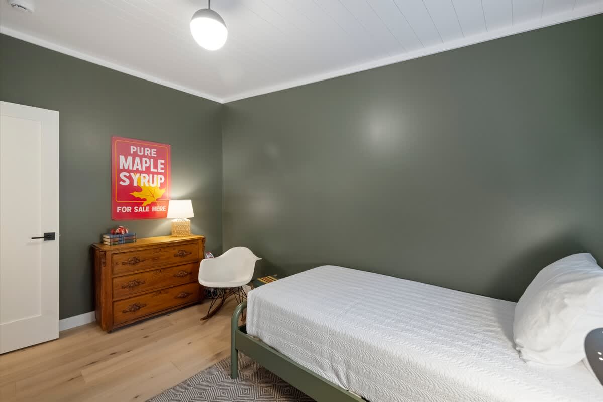 A cozy bedroom featuring a single bed with white bedding, a wooden dresser with a lamp, and a white rocking chair, all set against a dark green wall with light hardwood floors.