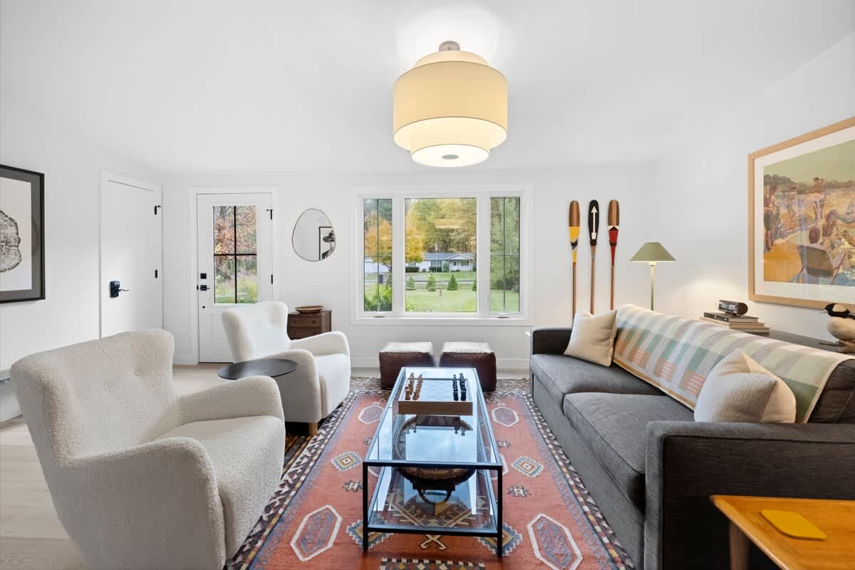 Bright living room with white walls and light wood floors, featuring a dark gray sofa and two white upholstered armchairs. A glass coffee table with a chess set is central, with a large window and a glass-paned door leading outside.