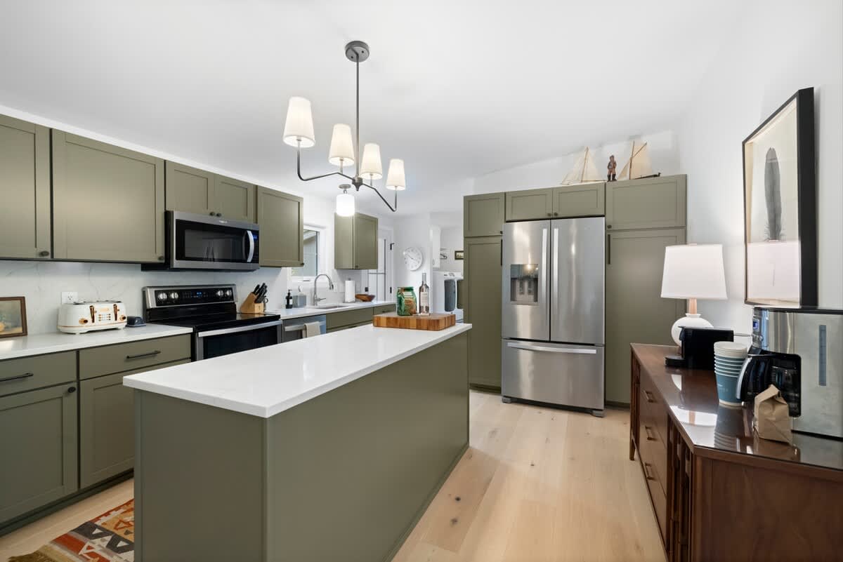 Modern kitchen featuring olive green shaker cabinets, white quartz countertops, stainless steel appliances, and a central island with a white top. A contemporary chandelier hangs above the island.