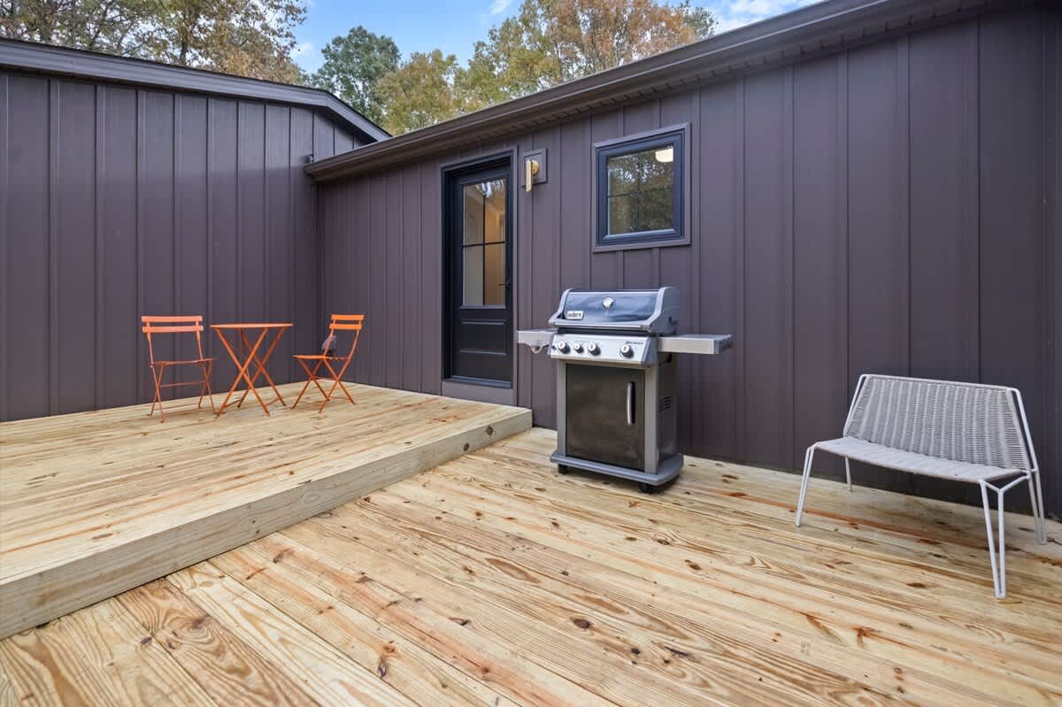 A section of the wooden deck featuring a stainless steel BBQ grill, a small orange bistro set, and a white woven chair, set against the dark brown board-and-batten siding of the house.