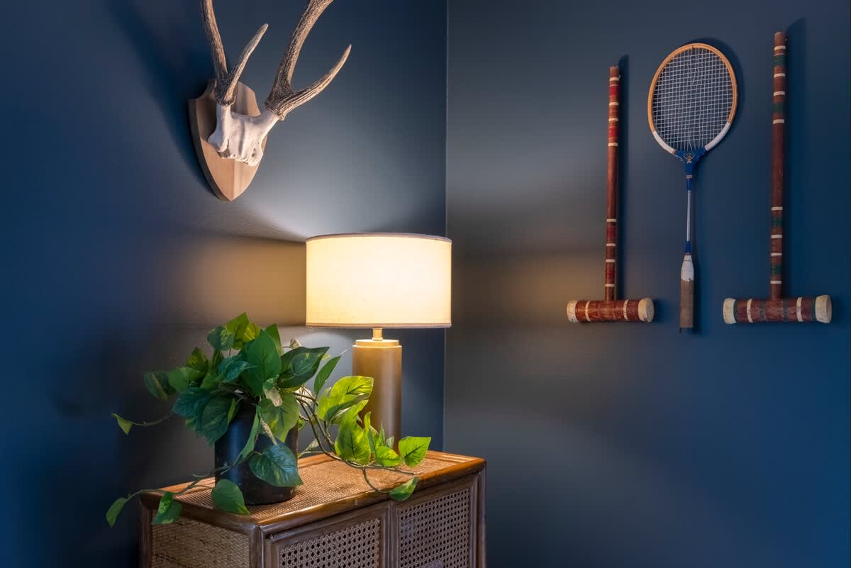 A detailed corner of the living room featuring dark blue walls, a rattan cabinet with a table lamp and a potted plant, and decorative deer antlers mounted above. Vintage croquet mallets adorn the wall.