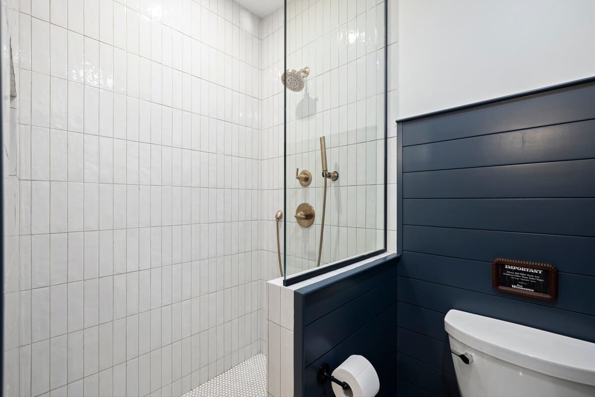 Modern walk-in shower with white vertical subway tiles and a glass partition, adjacent to a toilet area with dark shiplap walls. The shower features brass fixtures and a small tiled niche.