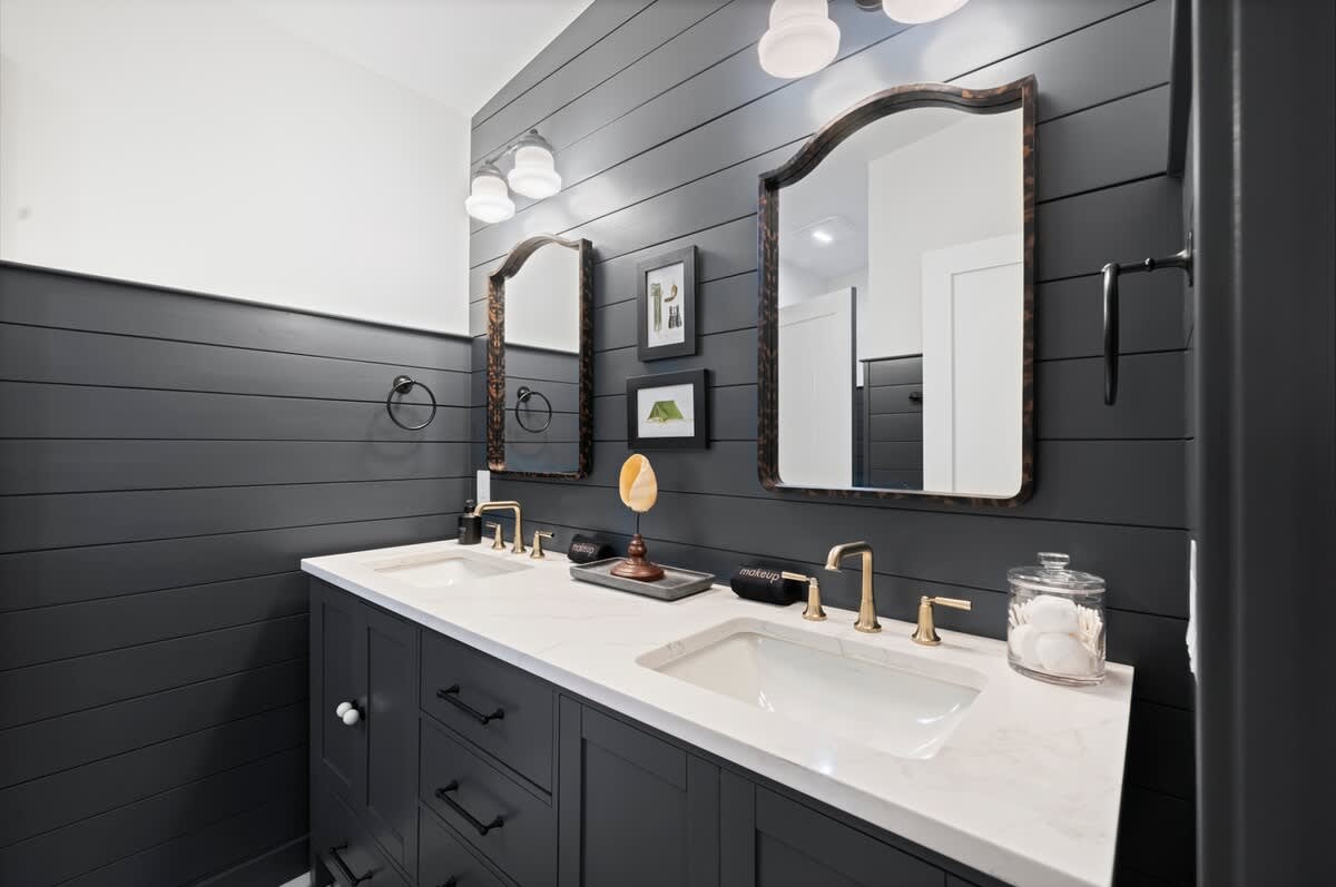Wide shot of a modern bathroom with dark shiplap walls, a double vanity with white quartz countertop, brass faucets, and two ornate mirrors. The room has white upper walls and a dark vanity cabinet.