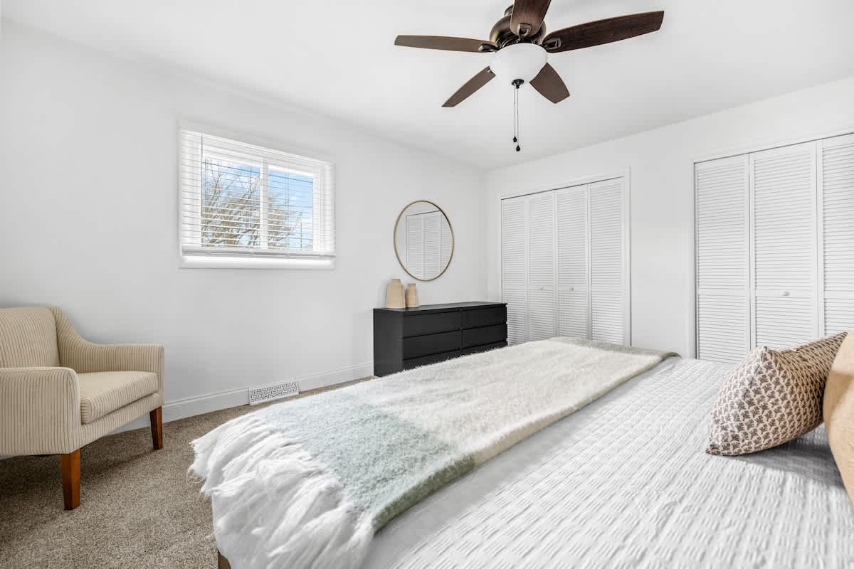 Another view of the bedroom, showing a cream armchair in the corner, a black dresser with a round mirror above it, and two windows with white blinds. The room has carpeted floors and a ceiling fan.