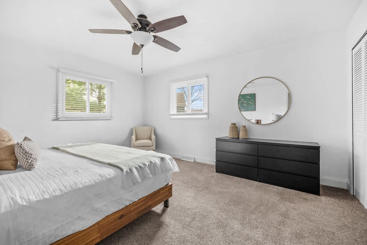 A wide shot of the bedroom, featuring the wooden bed, black dresser with a mirror, and an armchair, all against white walls with two windows providing natural light. The room is carpeted and has a ceiling fan.