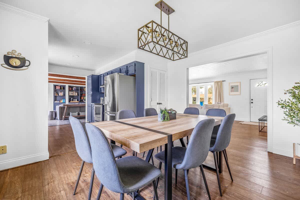 A modern dining room features a light wood rectangular table with a dark stripe, surrounded by six gray upholstered chairs, under a geometric gold chandelier, with a blue kitchen visible in the background.